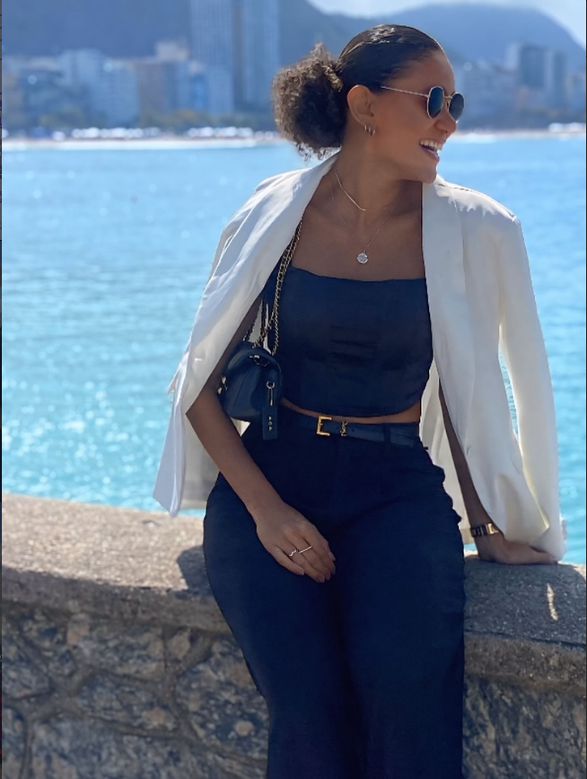 Psicóloga Adriana Andrade sentada elegantemente à beira-mar com blazer branco, sorrindo de perfil